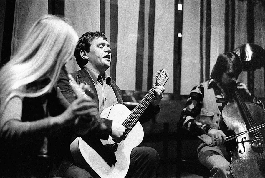 Fred Åkerström sjunger med gitarr på scen, mellan en flöjtist och en cellist.